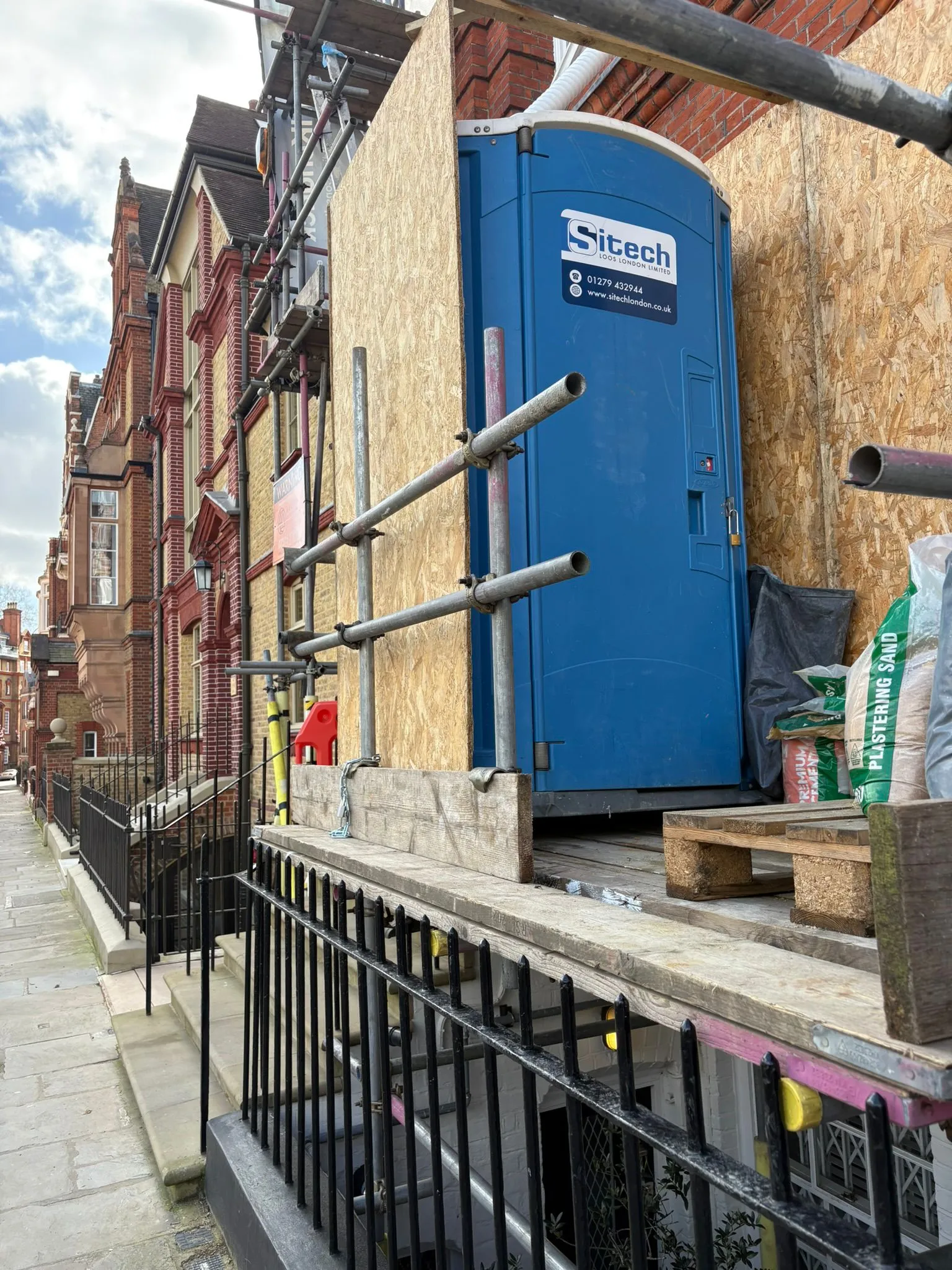 Close-up of a Sitech Loos London Maxim 3000 chemical toilet mounted on a scaffold deck against a London building facade