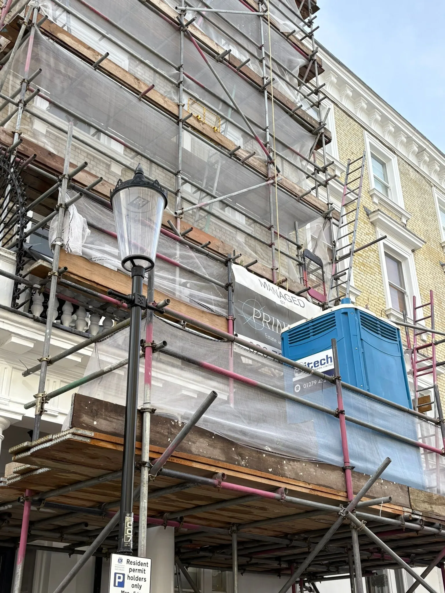 A blue Sitech Loos London Maxim 3000 portable toilet mounted high on scaffolding outside a London townhouse, viewed from street level