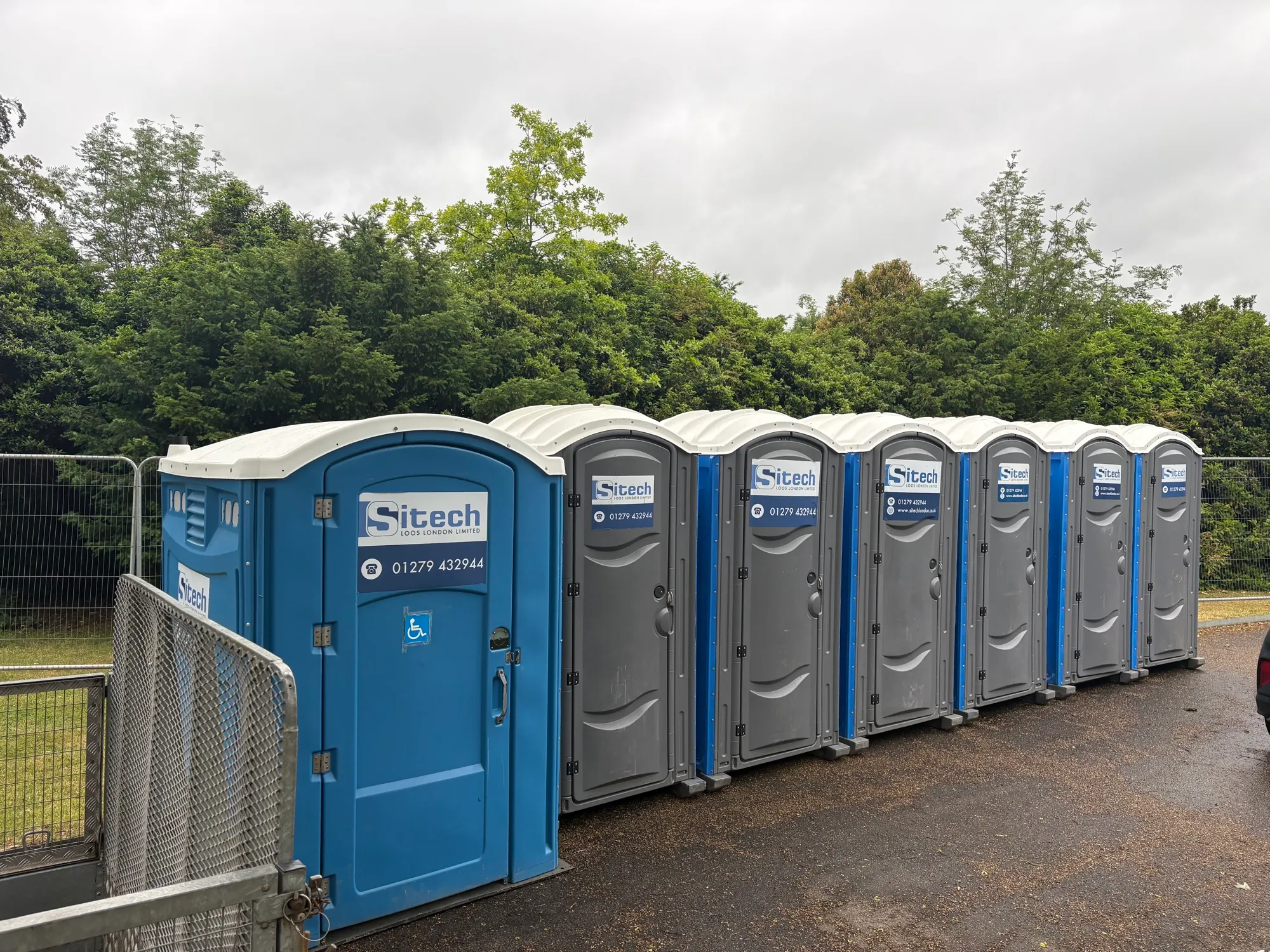 A Sitech Loos London disabled access portable toilet at the end of a row of six event toilets on tarmac at the BBC Antiques Roadshow, with the mesh gate of a trailer in the foreground